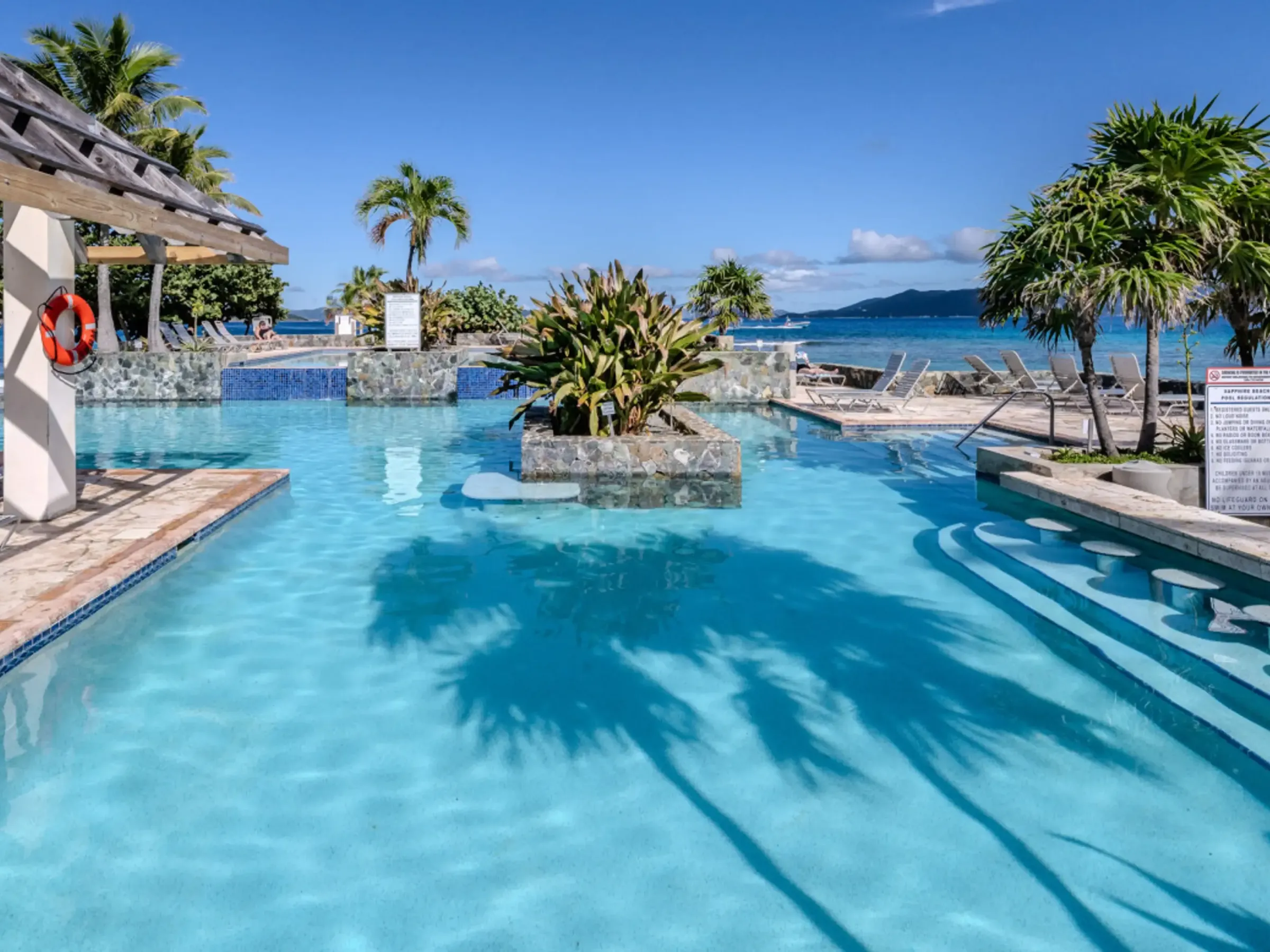 Tropical pool with palm trees, ocean view, and lounge chairs under clear sky.