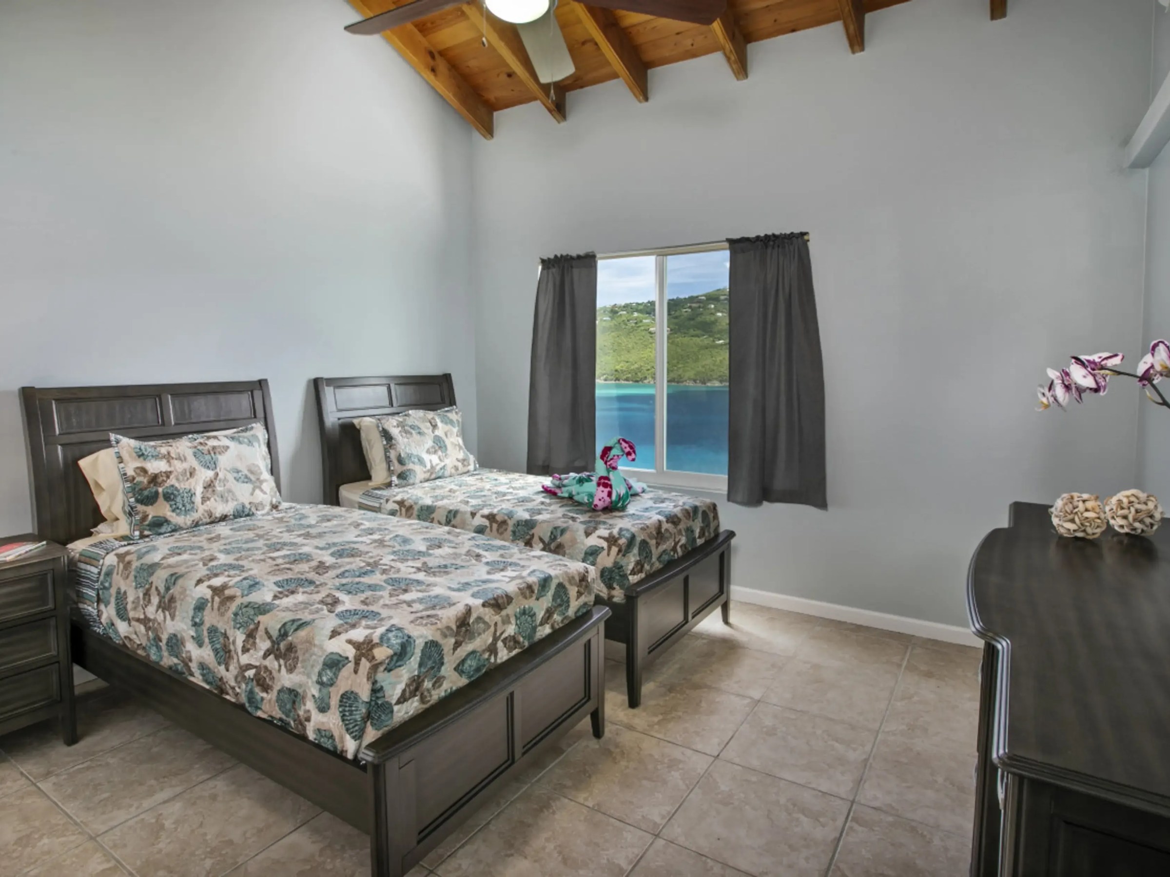 Bedroom with two twin beds, floral bedding, and a window with a lake view, ceiling fan, and wooden beams.