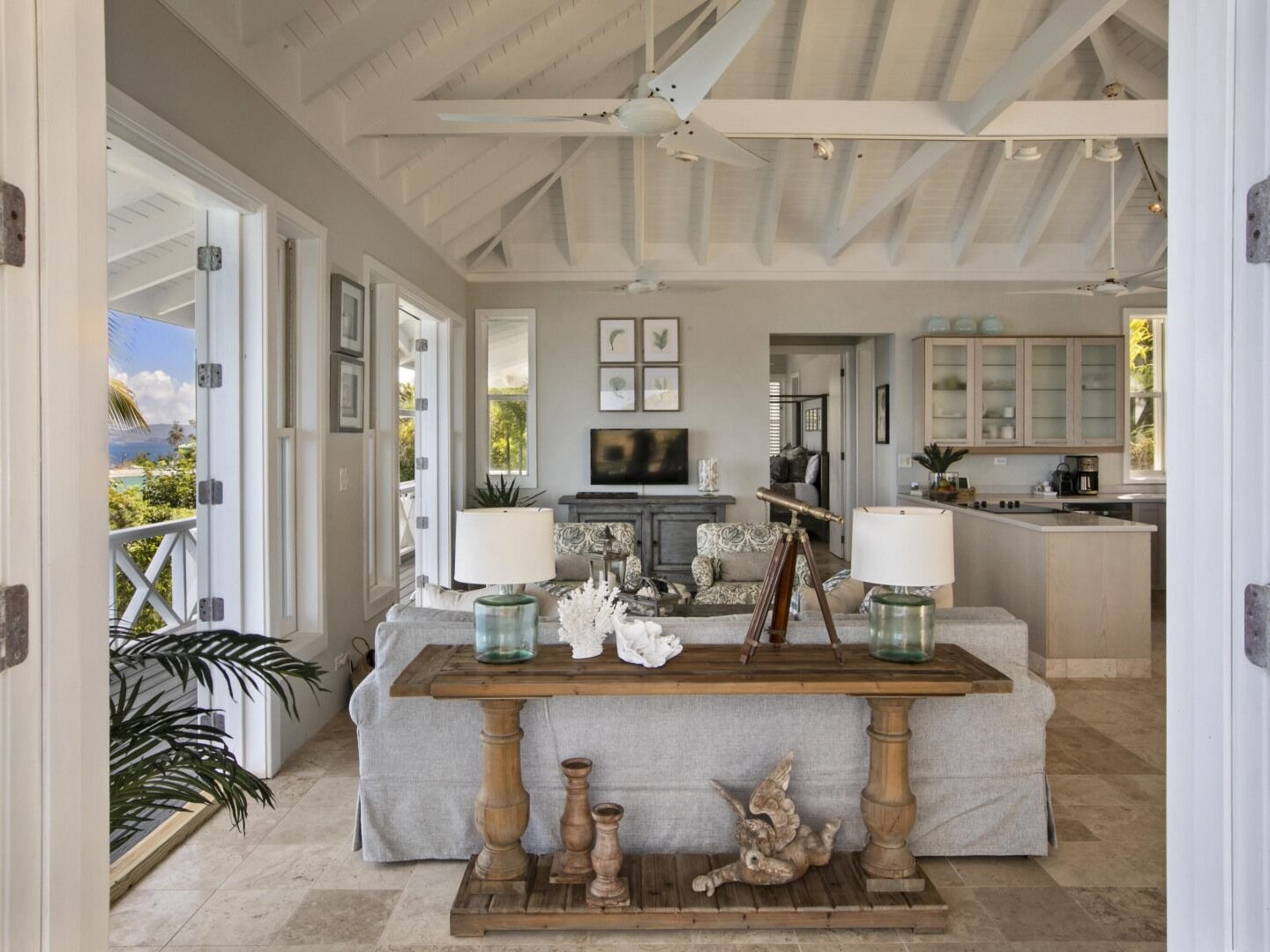 Bright living room with vaulted ceiling, sofa, table, decor items, and open doors revealing tropical view.