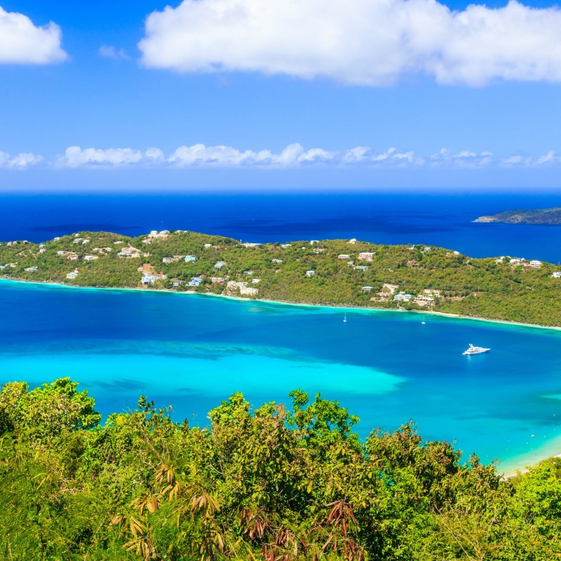 Aerial view of a tropical bay with turquoise water and green hills under a blue sky with scattered clouds.