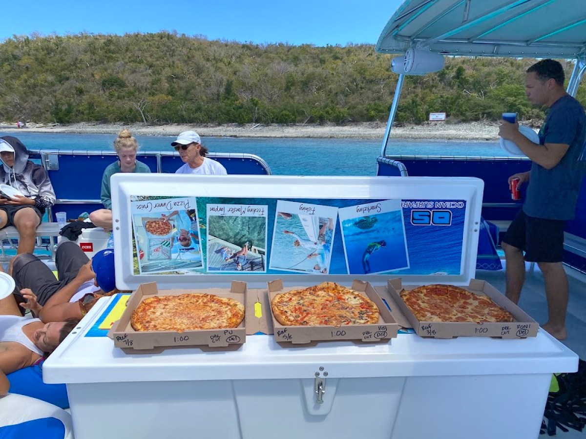Open pizza boxes on a boat with people and scenic ocean view in the background.