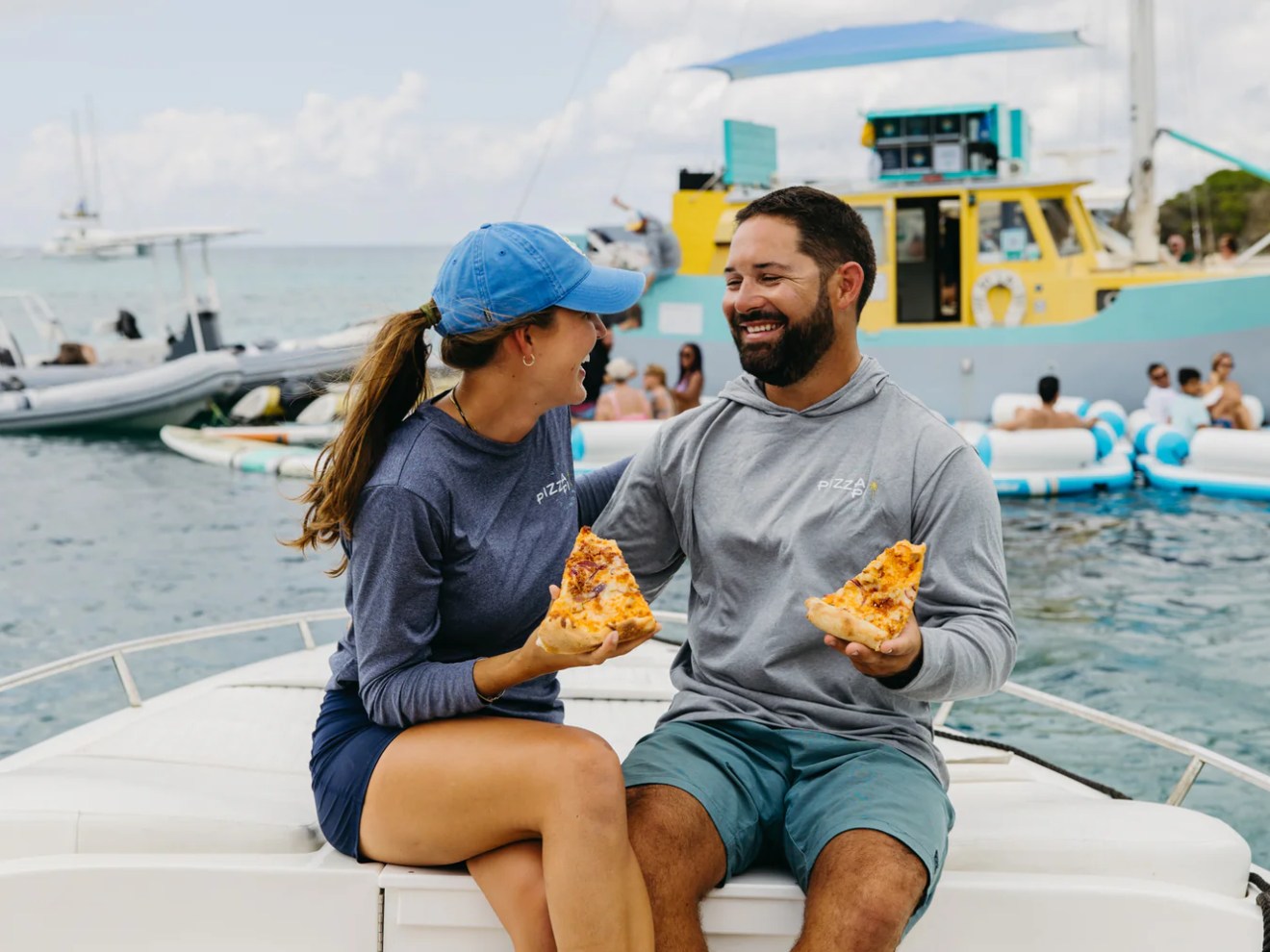Two people sitting on a boat, smiling and holding slices of pizza, with other boats and a barge in the background.